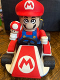 Toy figure of a character in a red hat and blue outfit with a mushroom and kart on a wooden surface.