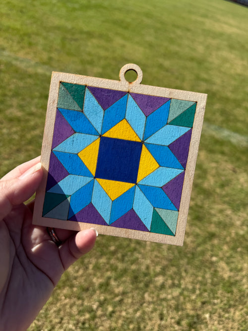Colorful barn quilt square ornament held in a female hand on a green background. 