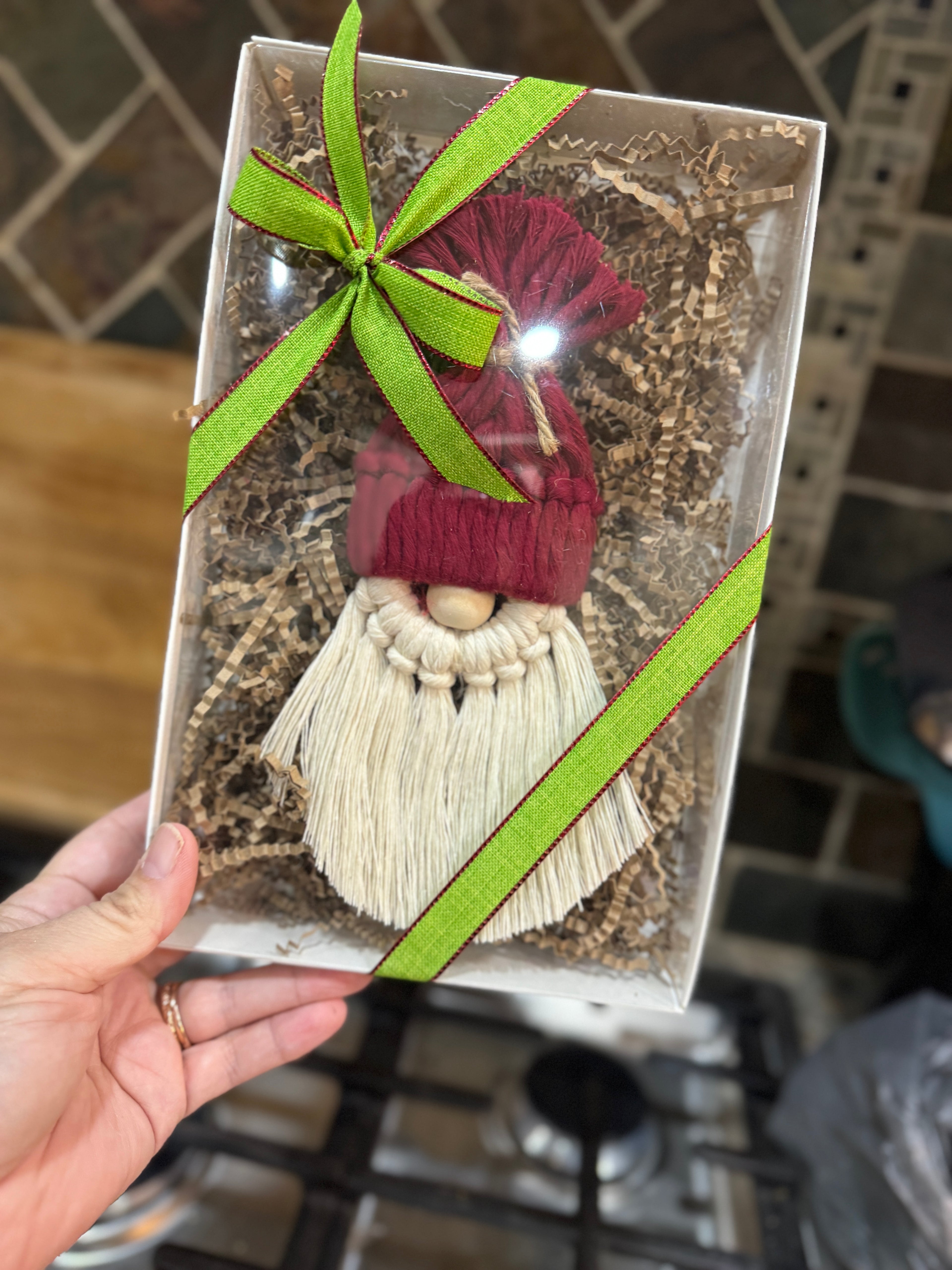 Decorative gnome figure in a clear box with a green ribbon, held by a hand on a tiled kitchen counter.
