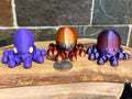 Three octopus-shaped figurines in purple, brown, and red on a wooden surface with a coin for scale.